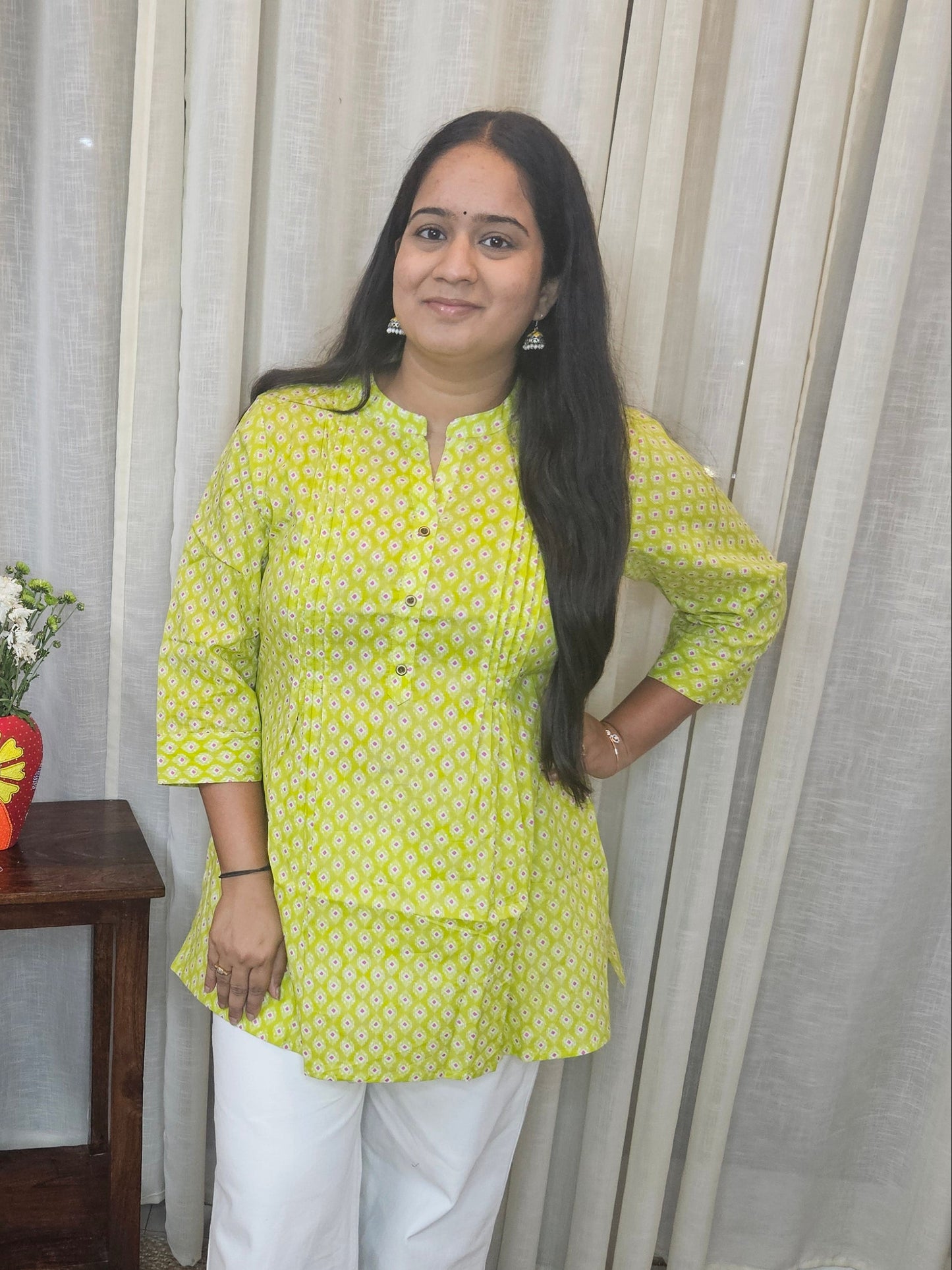 Woman in a yellow top and white pants standing in a room with curtains and a small table.