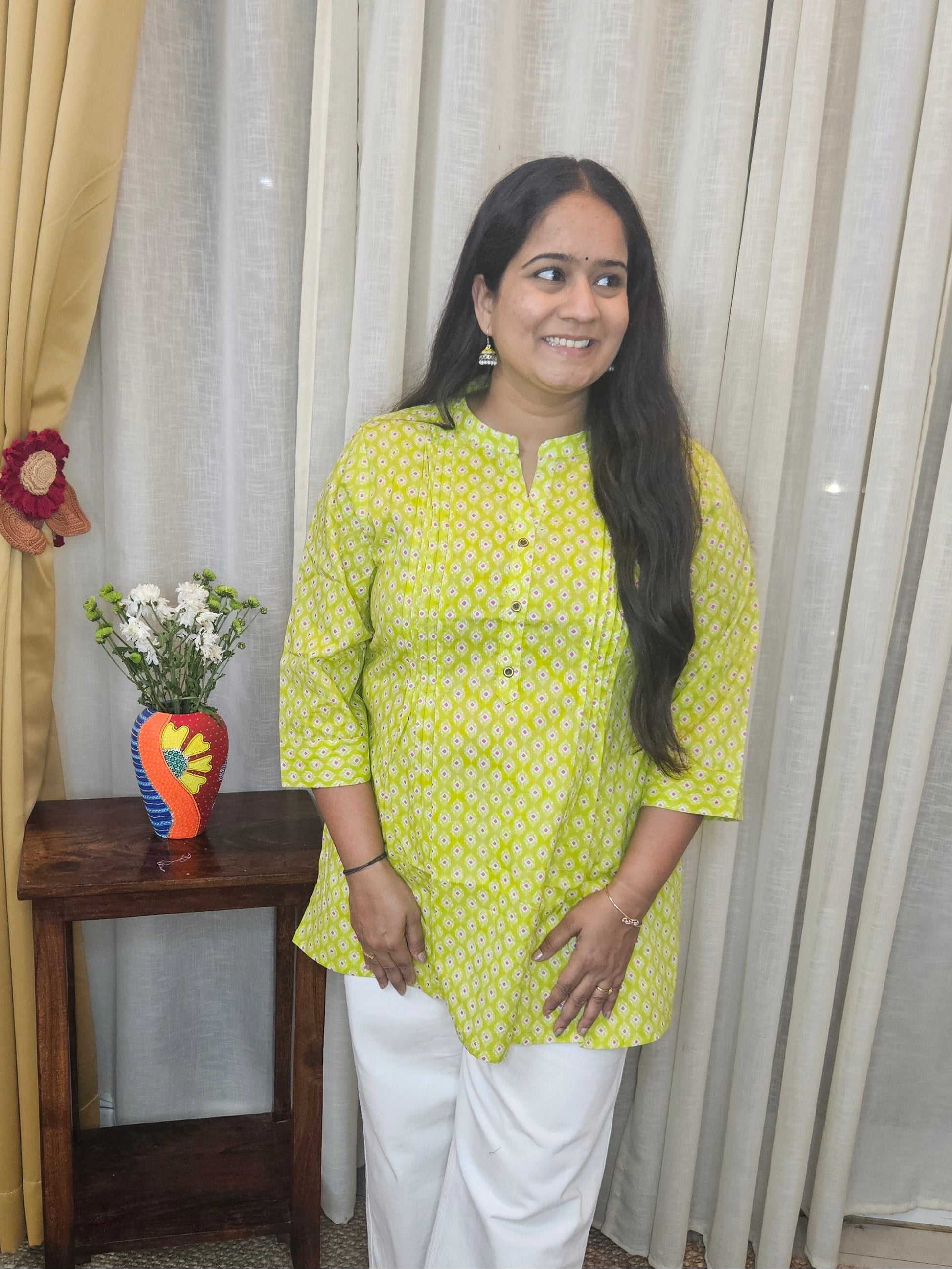 Woman in a yellow top and white pants standing in front of a curtain with a small table and decorative items.
