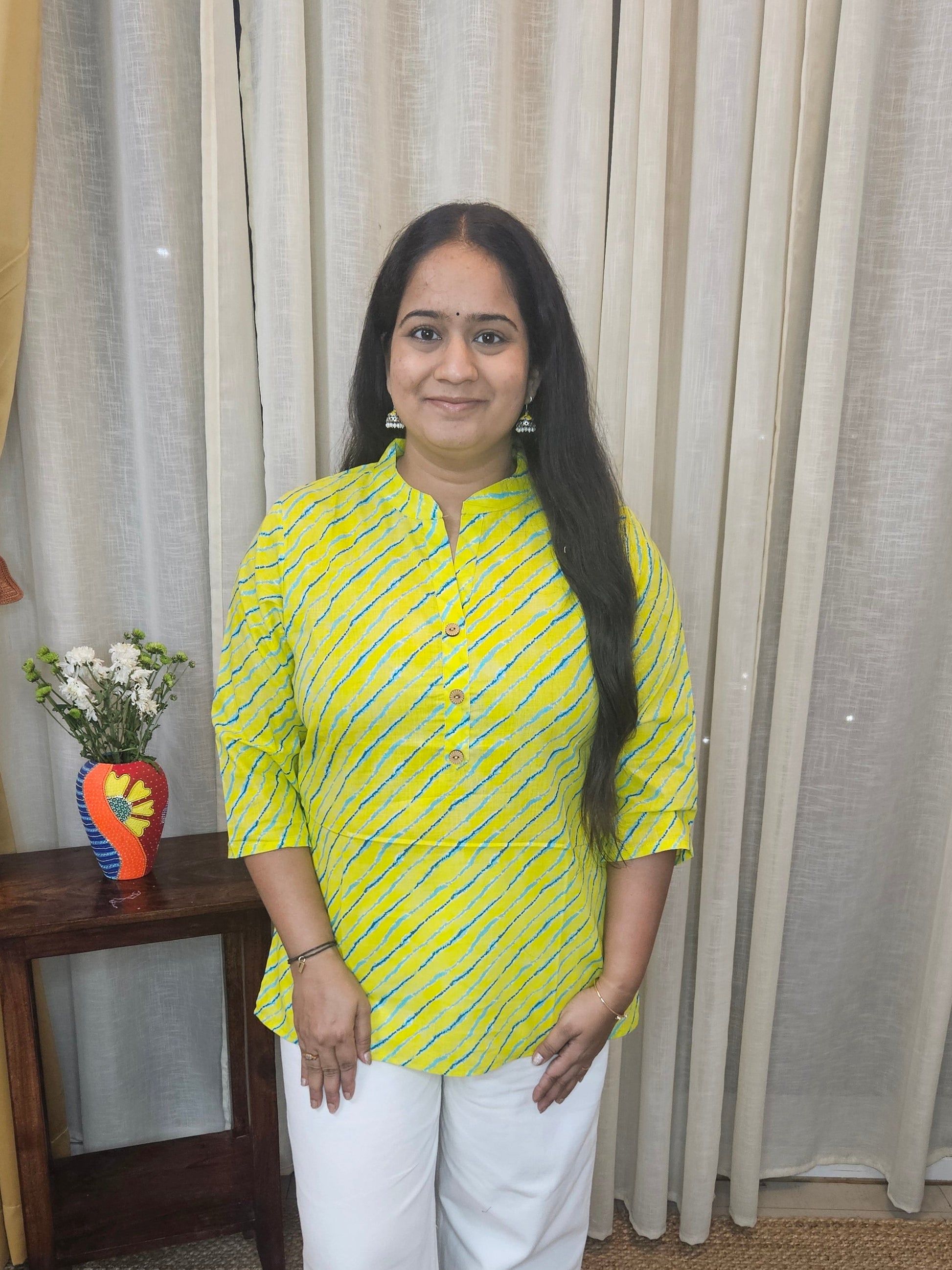 Woman in a yellow top and white pants standing in front of a curtain.