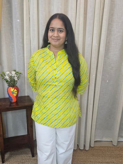 Woman in a yellow top and white pants standing in a room with curtains and a small table.
