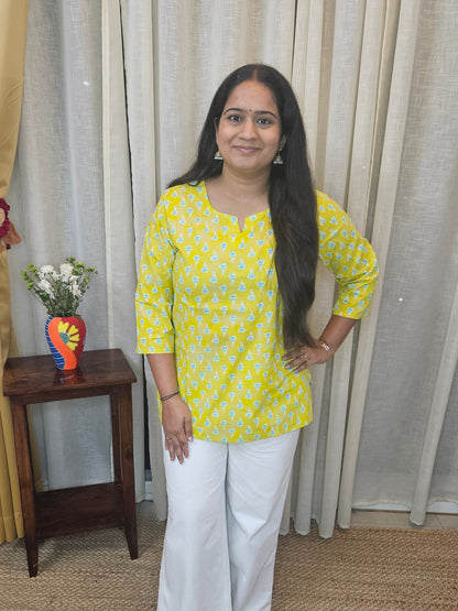 Woman in yellow top and white pants standing on a staircase with curtains and a small table in the background.