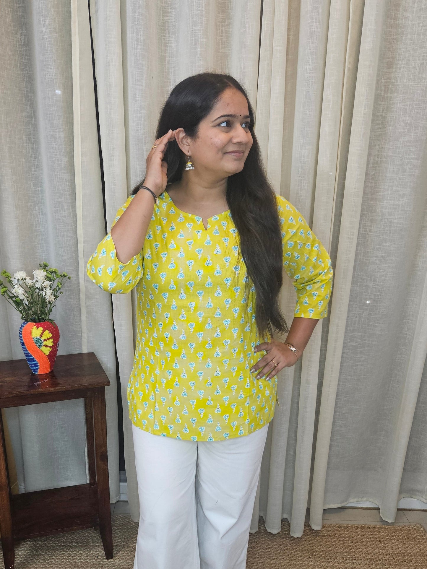 Woman in yellow top and white pants standing in a room with curtains and a small table.