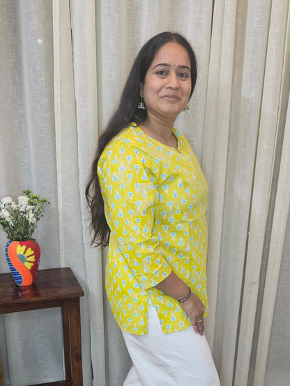 Woman standing on a staircase wearing a yellow top and white pants, with a decorative flower arrangement on a small table.